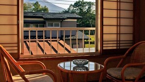 A room with a view of a house and a mountain. There are two wooden chairs and a small table with a glass object on it. - Oogiyama Ryokan (Fujikawaguchiko)