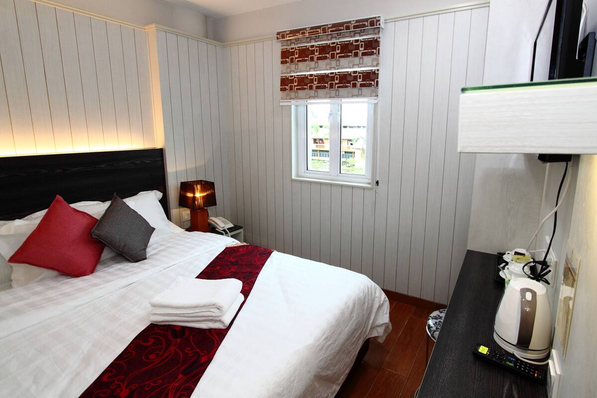 A neatly made bed with white bedding and red and gray pillows, a bedside table with a lamp, a window with a view, and a kettle on the table.