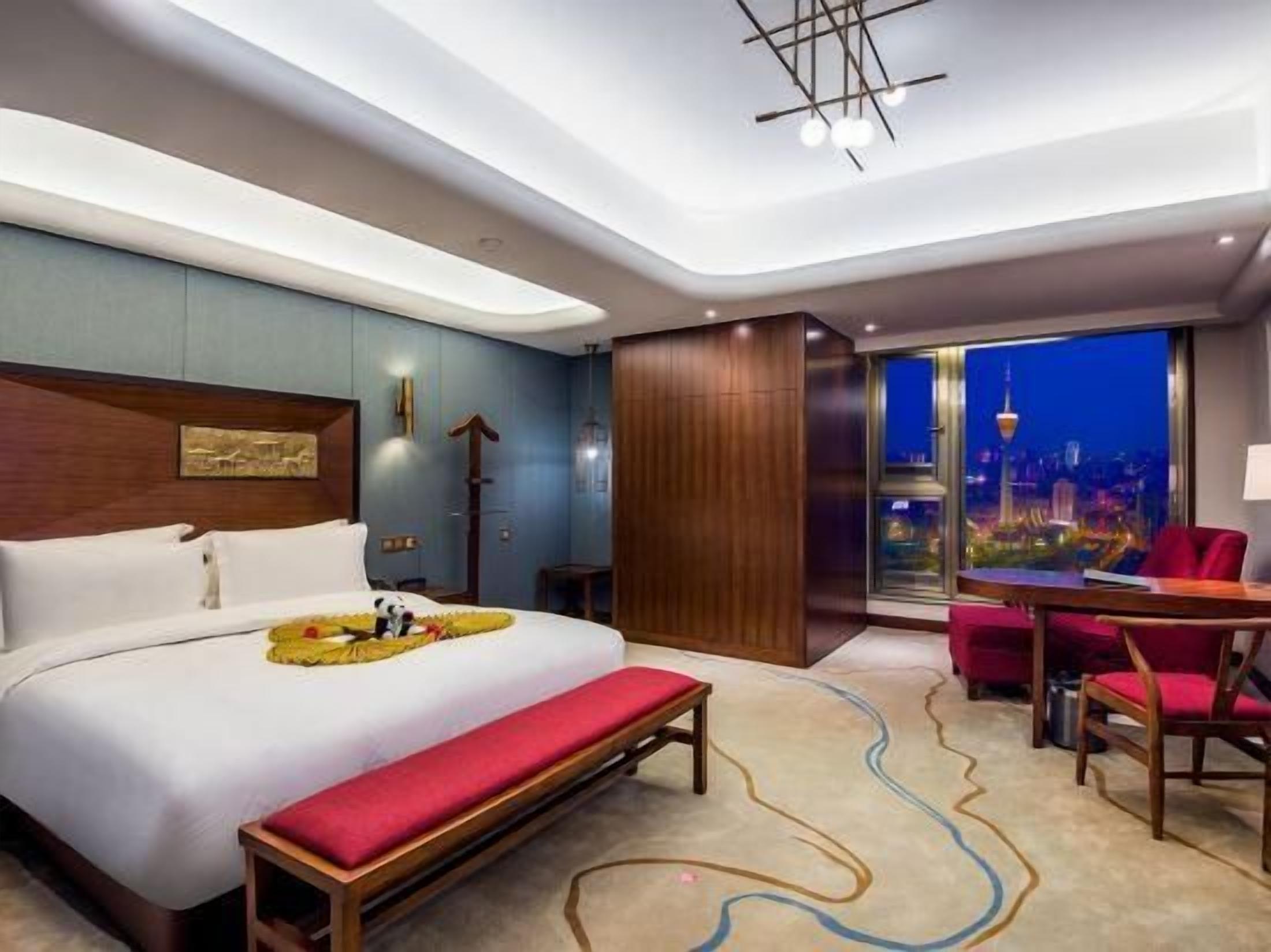 A modern hotel room with a large bed, a bench, a desk, and a view of a cityscape.