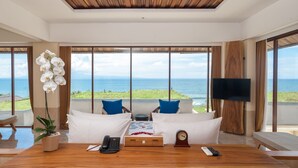 A modern hotel room with a wooden ceiling, large windows with ocean views, a wooden table with a telephone, remote, and a clock. - Komaneka at Keramas Beach (Gianyar)