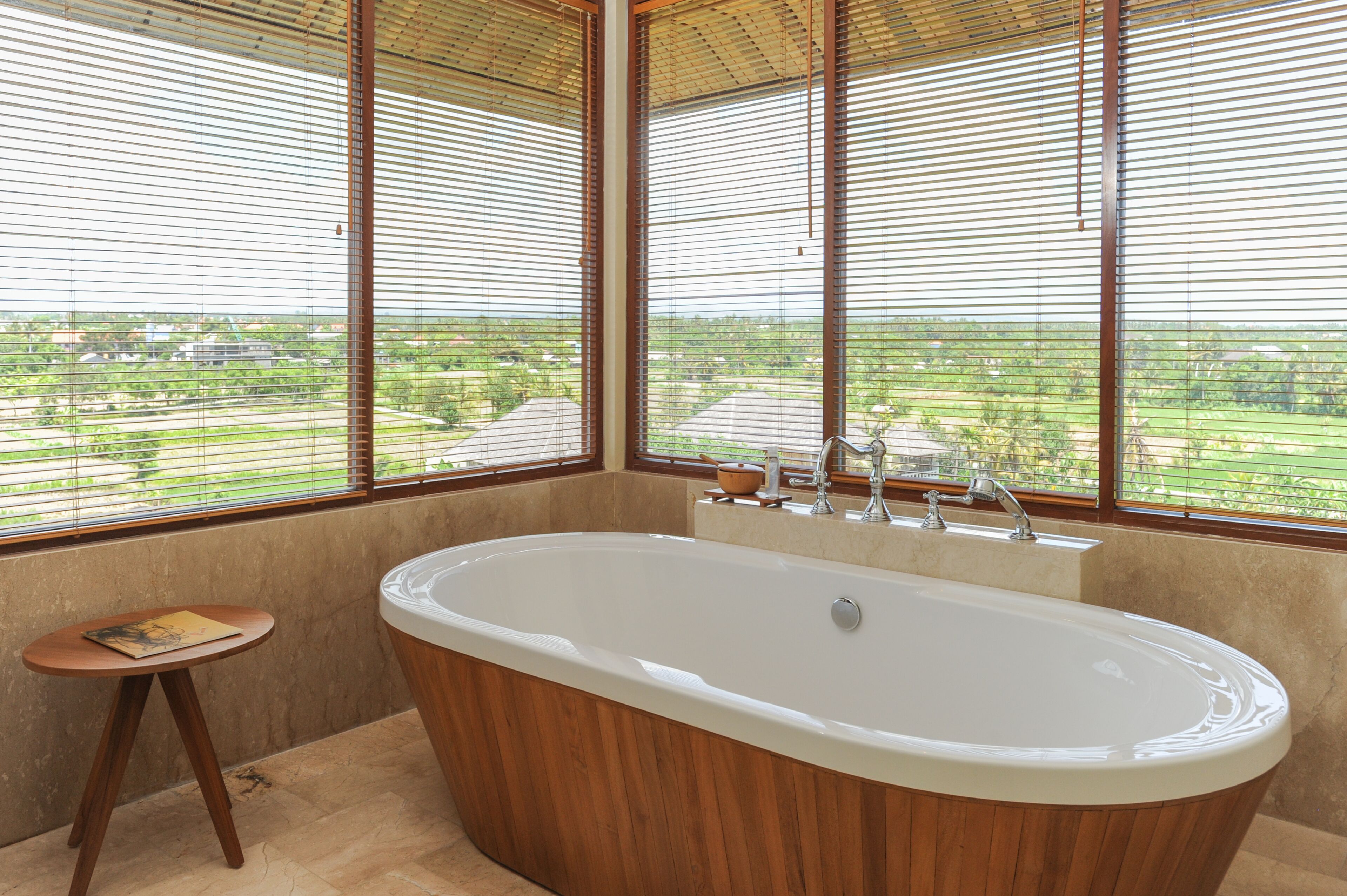 Ein modernes Badezimmer mit einer freistehenden Badewanne, großen Fenstern mit Jalousien und einem kleinen Holzschemel.