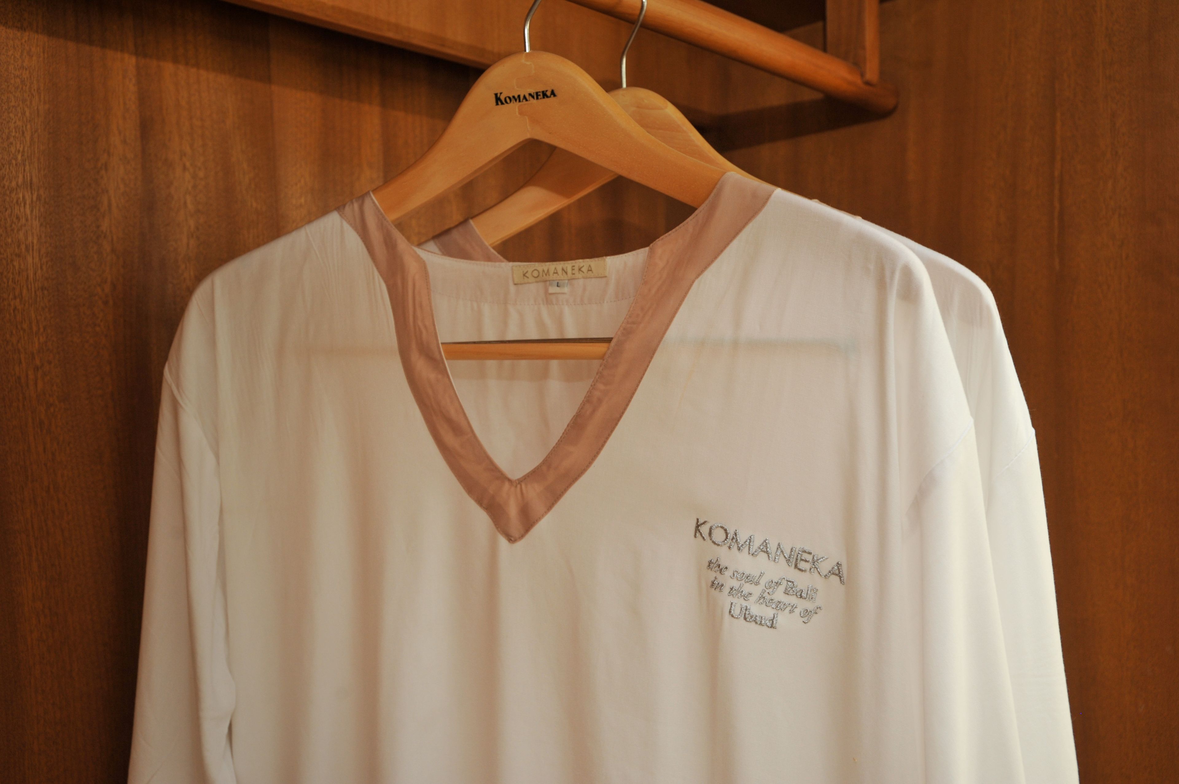 Una camicia bianca con scollo a V e il logo Komaneka sul lato sinistro, appesa a un appendiabiti in legno.