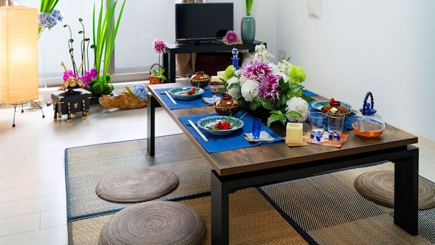 A dining area with a wooden table set for a meal, surrounded by woven mats and a TV in the background.