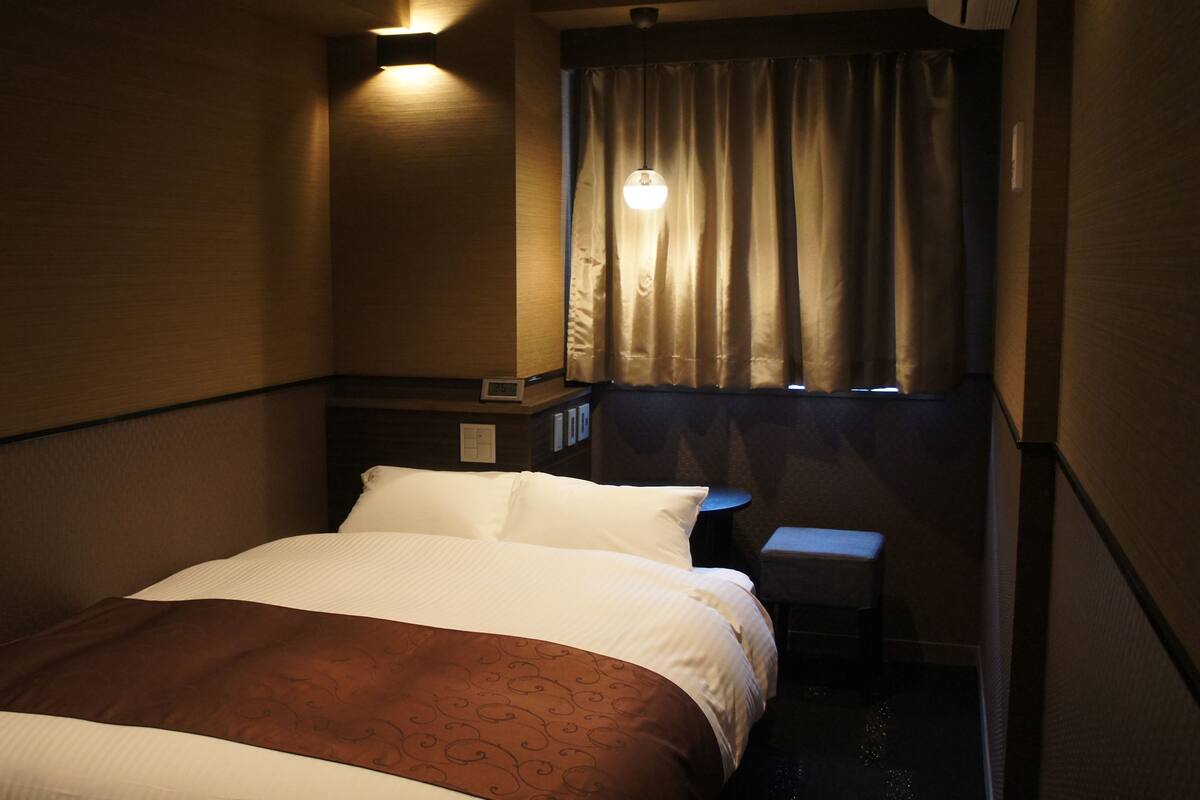 Una habitación de hotel con cama, mesita de noche, una pequeña mesa y una ventana con cortinas.
