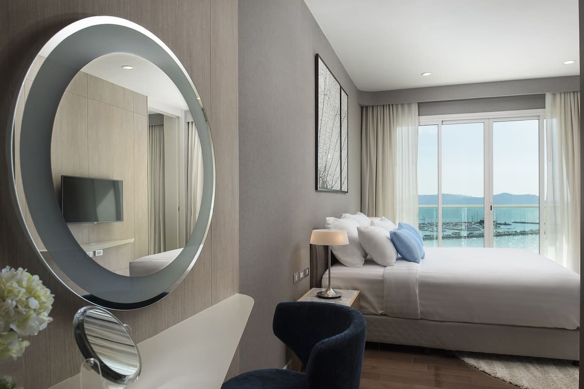 Ein modernes Hotelzimmer mit einem großen Bett, einer Sitzecke mit einem Sessel, einem runden Spiegel, einem Flachbildfernseher und Meerblick durch große Fenster.