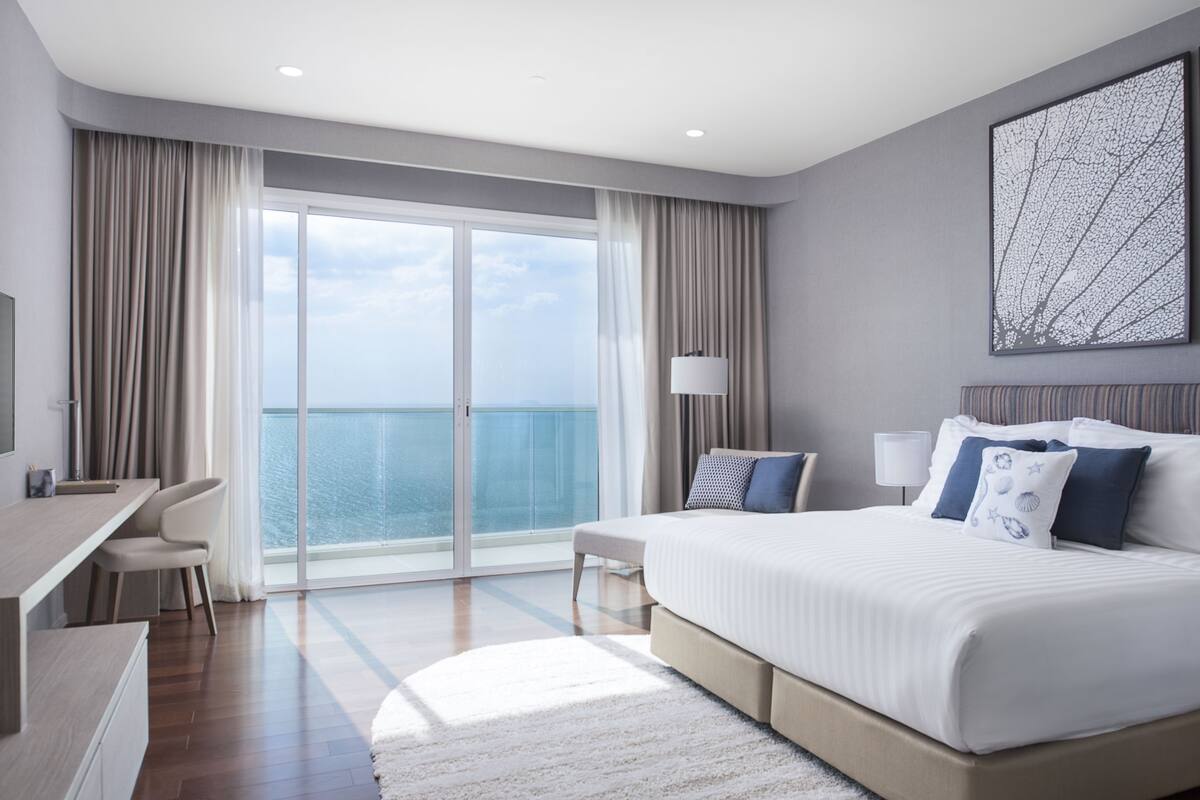Ein modernes Hotelzimmer mit einem großen Bett, einem Schreibtisch und Meerblick durch raumhohe Fenster.