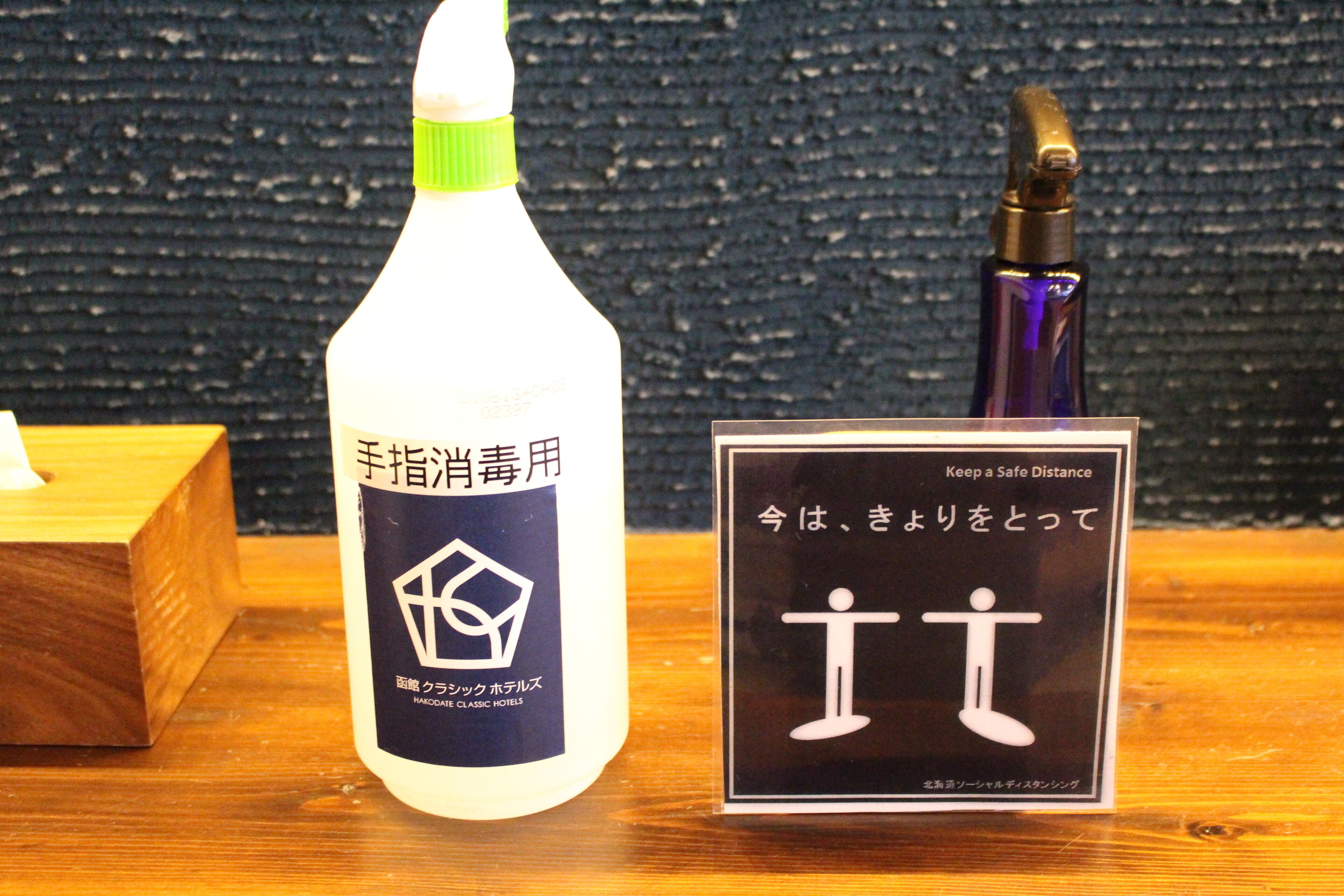 Une bouteille de gel hydroalcoolique et un panneau avec du texte en japonais et un pictogramme indiquant qu’il faut respecter une distance de sécurité.