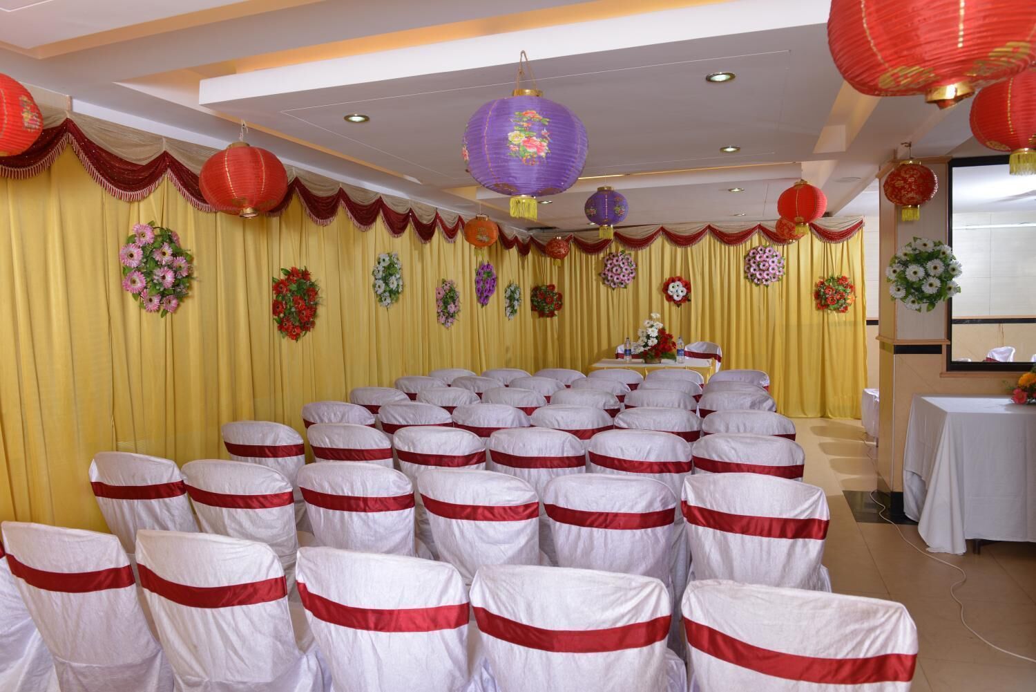 Un salón de banquetes con filas de sillas cubiertas con lazos blancos y rojos, cortinas amarillas y faroles rojos y morados.