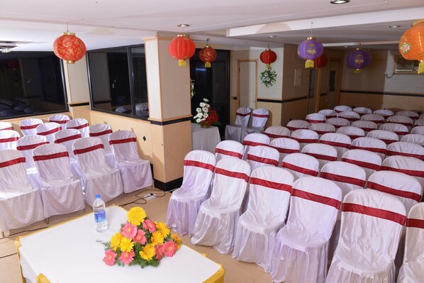 Un salón de conferencias con filas de sillas adornadas con cintas blancas y rojas, una mesa con un centro de mesa floral y faroles rojos y morados colgando del techo.