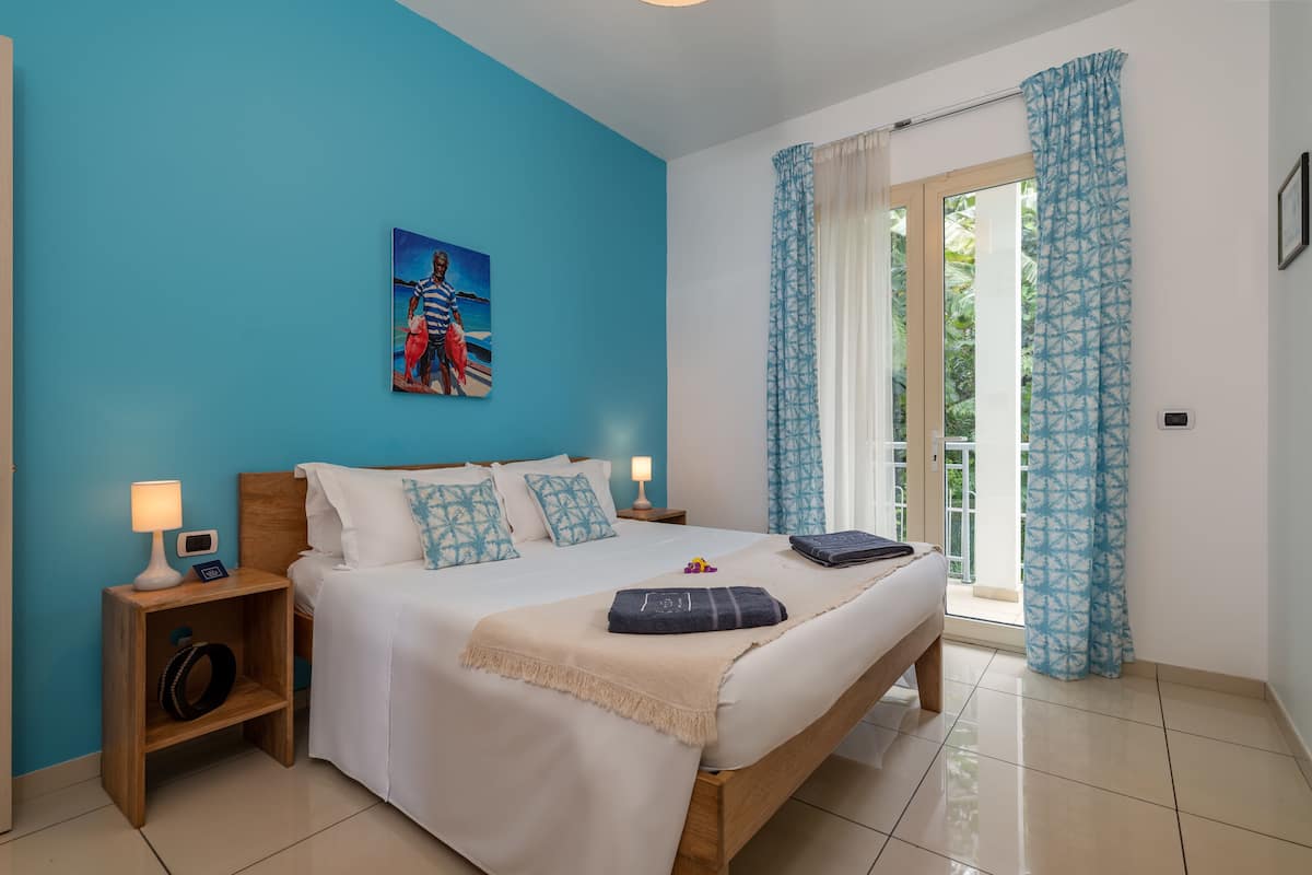 A bedroom with a bed, nightstands, a painting, and a view through a window with curtains.
