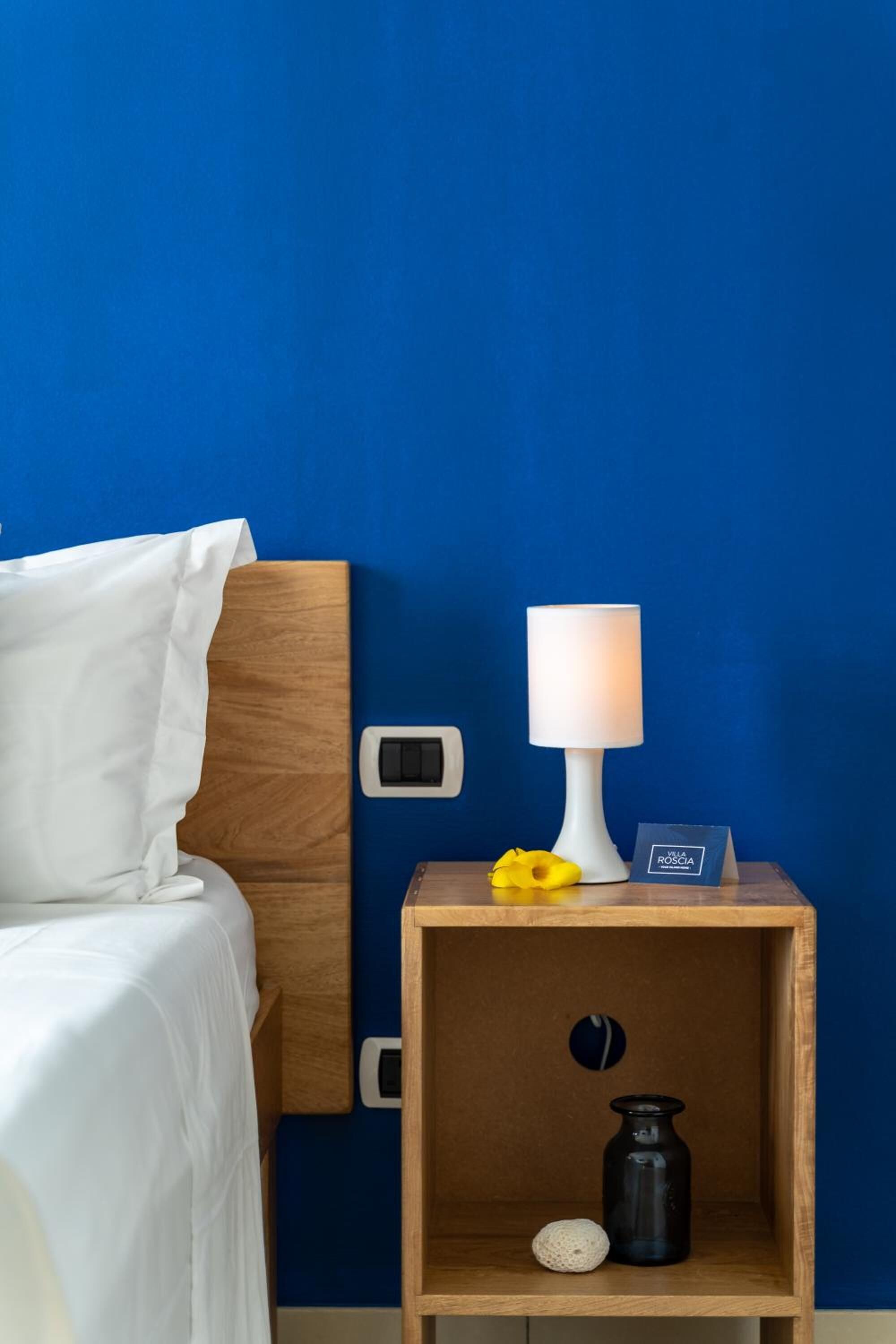 Una cama bien tendida con sábanas blancas, una mesita de noche de madera con una lámpara blanca, un pequeño florero y un adorno.