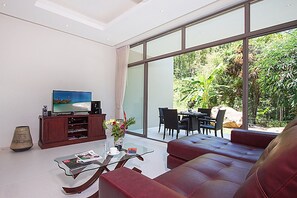 A modern living room with a large TV, comfortable seating, a glass coffee table, and a dining area with a view of greenery outside. - 2 Bedroom Pool Villa with Waterfall View SDV006-By Samui Dream Villas (Koh Samui)