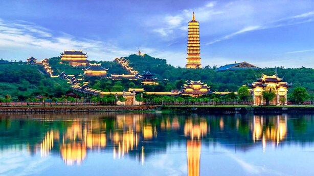 Una vista tranquila de un lago con arquitectura tradicional, incluyendo pagodas y una torre de estilo pagoda, reflejándose en el agua.