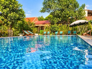A swimming pool with tiled edges, surrounded by greenery and a building with a red roof in the background.