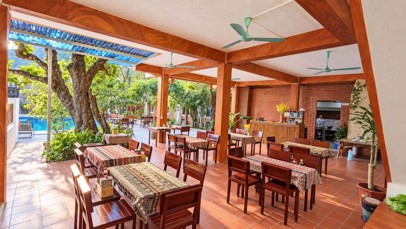 An outdoor dining area with wooden tables and chairs, a brick wall, and a view of a pool and trees.