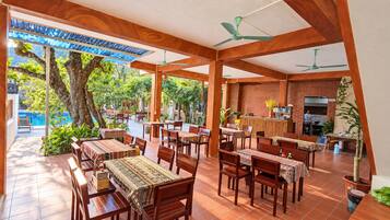 An outdoor dining area with wooden tables and chairs, a brick wall, and a view of a pool and trees.