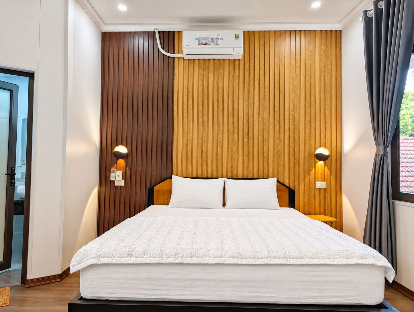 Quarto de hotel com cama, parede de painéis de madeira, ar condicionado e duas luminárias fixadas na parede.