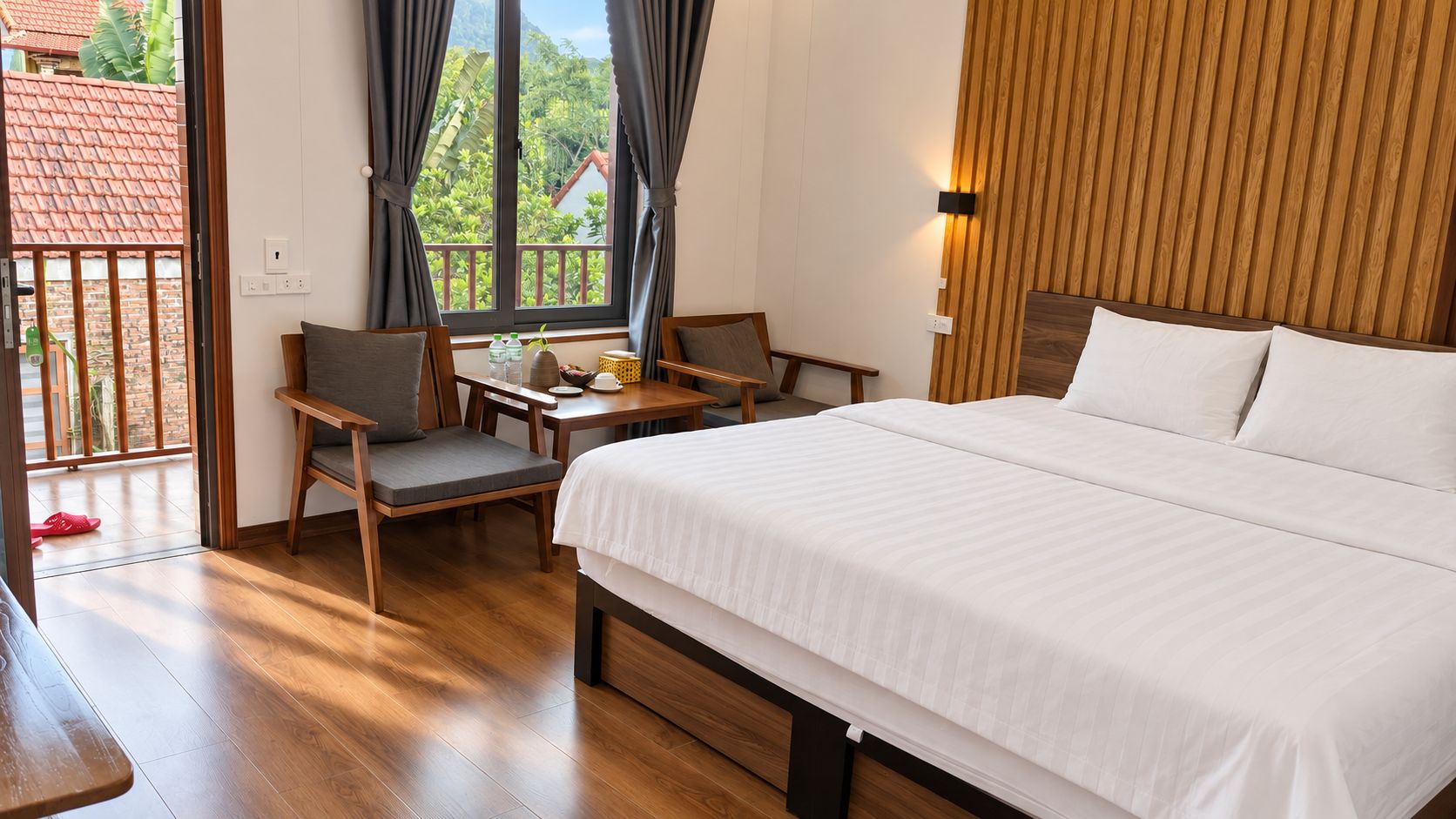 A hotel room with a bed, a desk, two chairs, a window with curtains, and a view of greenery outside.