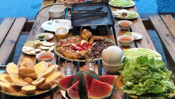 Mesa de madera con diversos alimentos y una parrilla, frente a una piscina.