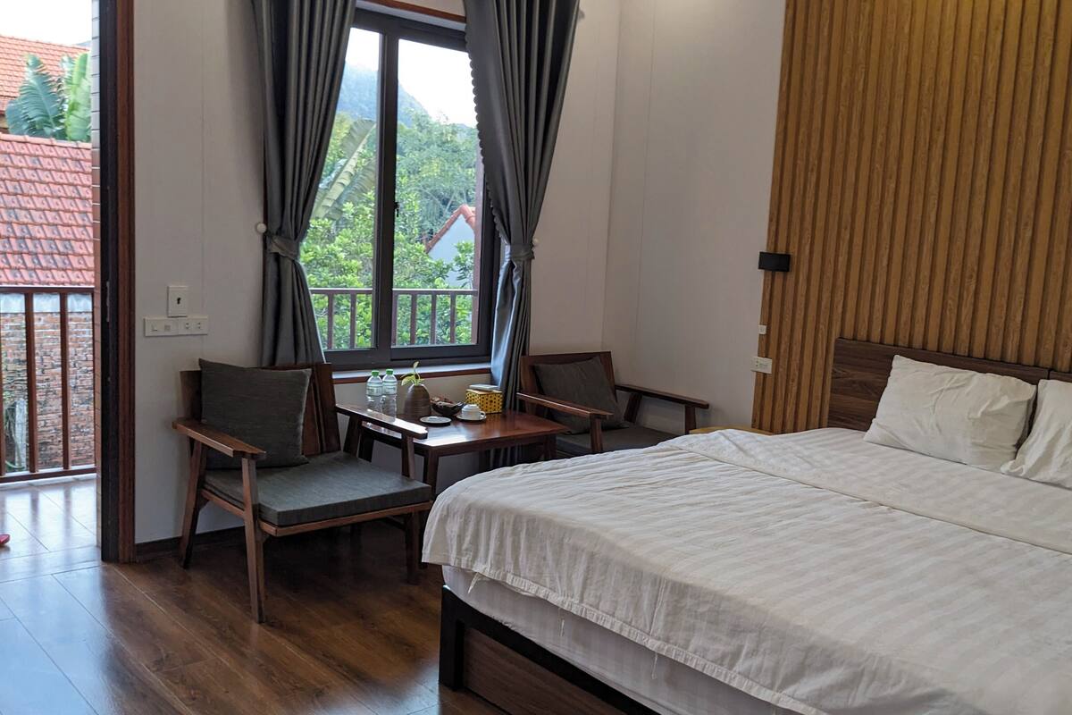 Une chambre d’hôtel avec un lit, deux chaises, une petite table avec quelques objets, une fenêtre avec des rideaux et une vue sur la verdure à l’extérieur.