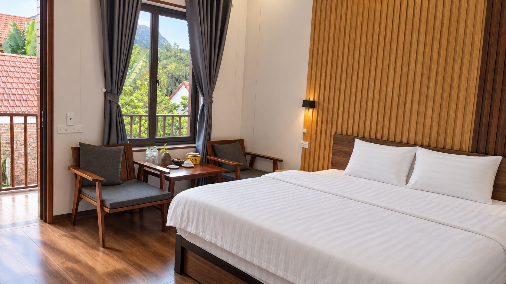 A hotel room with a bed, two chairs, a small table, and a view of greenery outside.