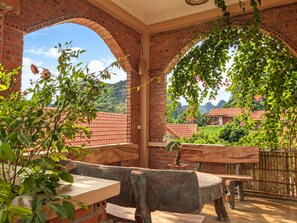 An outdoor patio with brick arches, wooden furniture, and a view of a green landscape with houses.