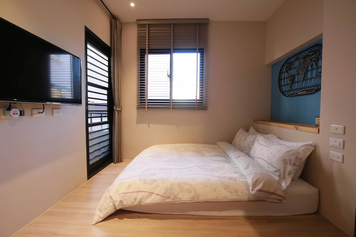 A bedroom with a bed, a flat-screen TV mounted on the wall, a window with blinds, and a world map decoration.