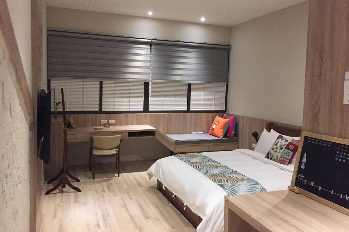 A bedroom with a bed, a desk, a chair, and a window with blinds.