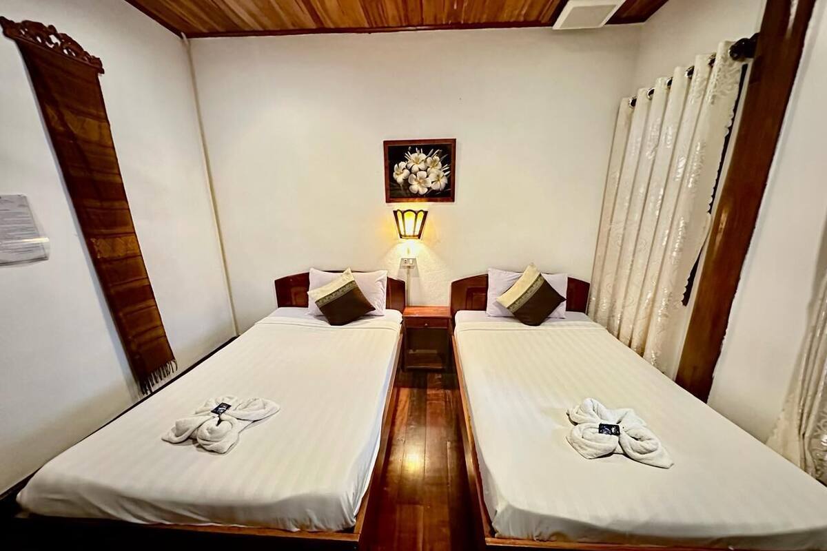 A hotel room with two single beds, a nightstand with a lamp, a framed floral artwork on the wall, and a wooden ceiling.
