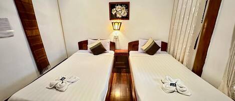 A hotel room with two single beds, a nightstand with a lamp, a framed floral artwork on the wall, and a wooden ceiling.