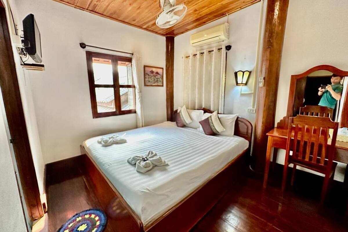 A hotel room with a wooden ceiling, a bed with white linens, a wooden chair, a desk, and a wall-mounted air conditioner.