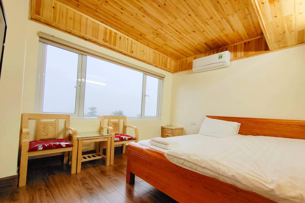 Quarto com teto de madeira, uma janela grande, cama com roupa de cama branca e escrivaninha de madeira com cadeiras.