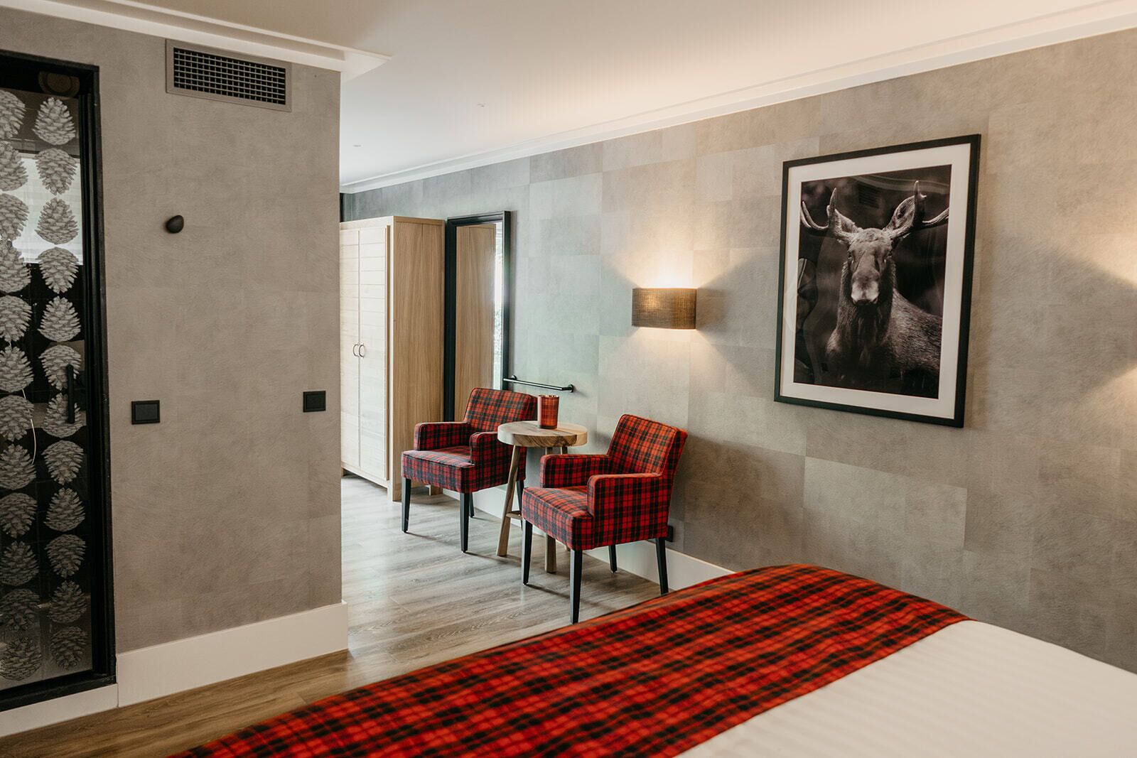 Une chambre d’hôtel avec un lit, un fauteuil à carreaux rouges, une petite table et un tableau encadré représentant un cerf, accroché au mur.