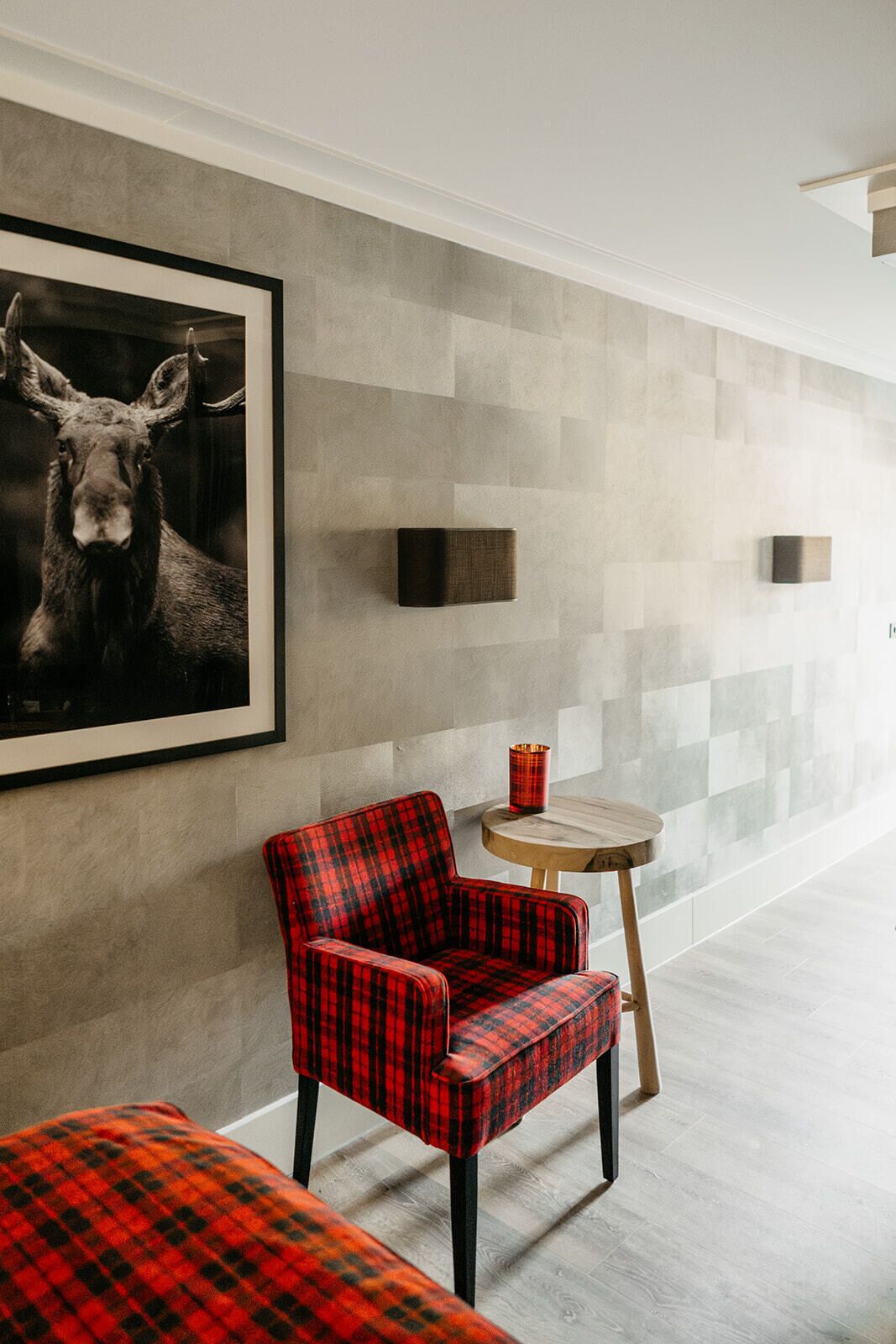 A modern hotel room with a large framed moose picture, a plaid chair, a small round table with a red candle, and a checkered blanket on the floor.