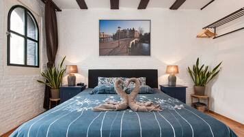 A bedroom with a large bed, a cityscape painting, and two bedside tables with lamps and plants.