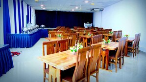 Ein Konferenzraum mit mehreren Holztischen und Stühlen, einer Bühne mit blauem Hintergrund und Deckenventilatoren.