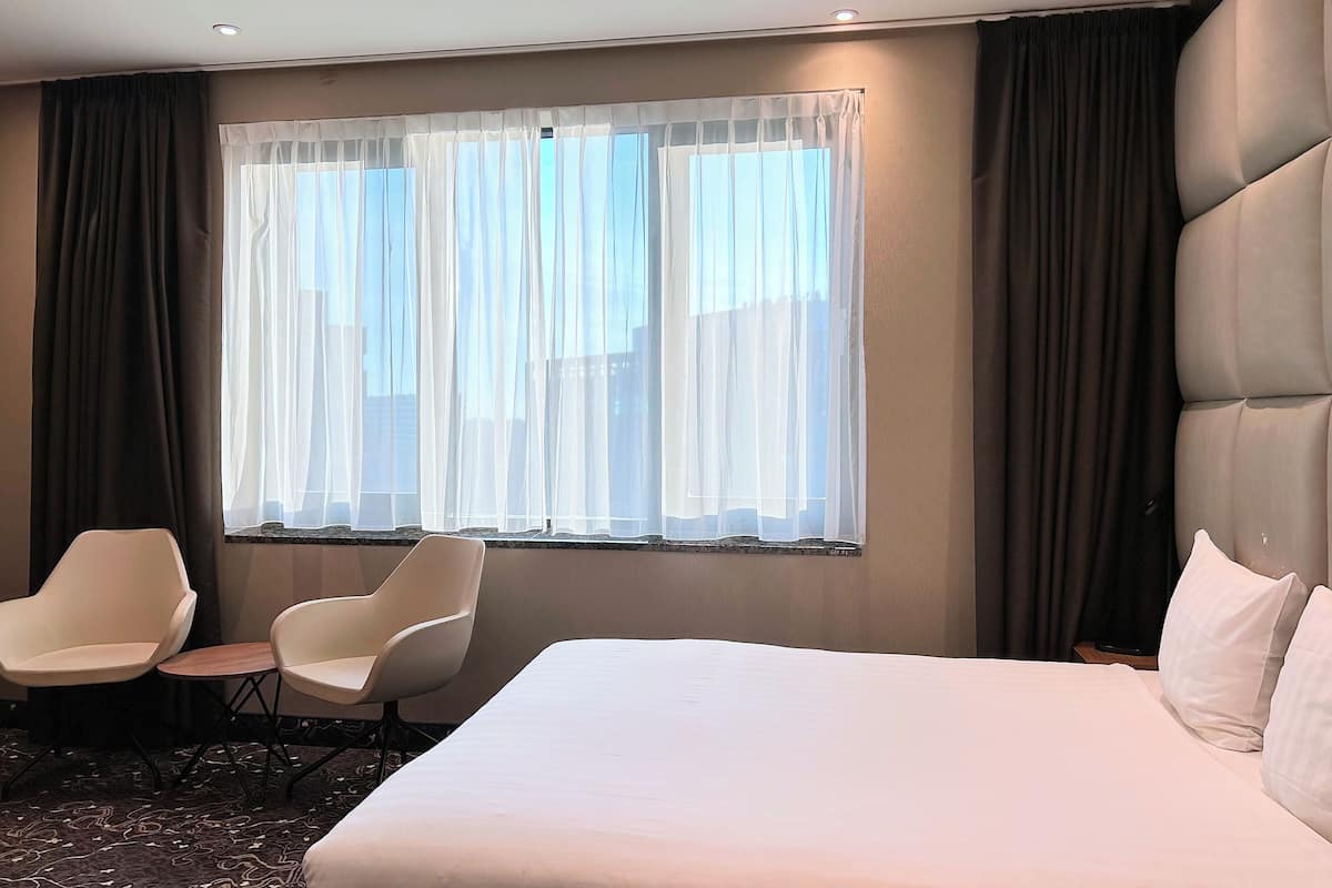 A hotel room with a large bed, two white chairs, a small table, and a large window with sheer curtains.