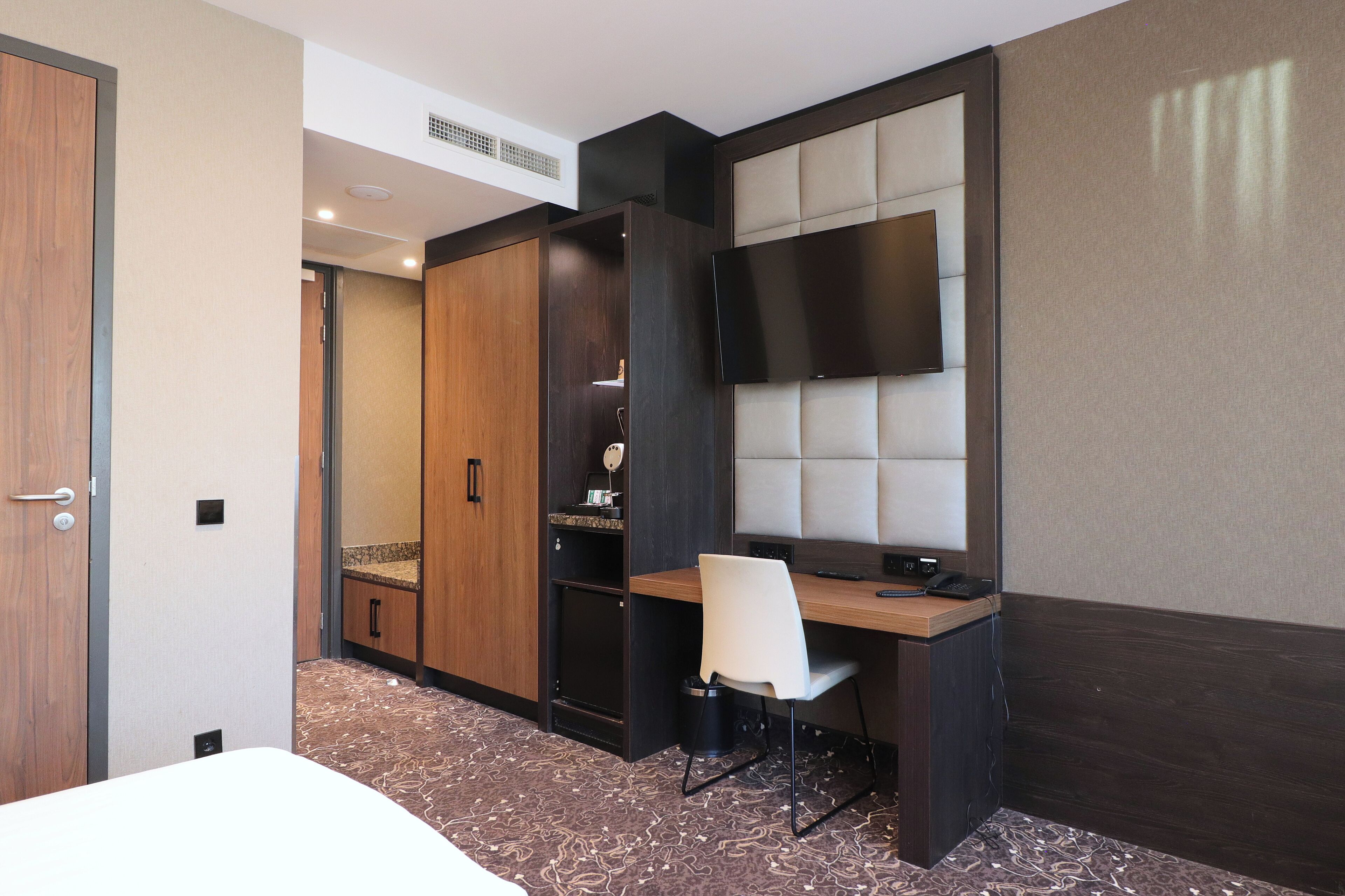 A hotel room with a bed, a desk with a chair, a wardrobe, a television mounted on the wall, and a patterned carpet.