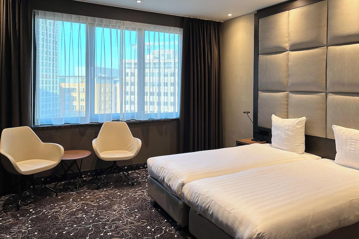 A hotel room with a large bed, two white chairs, a small table, and a view of the city through a large window with sheer curtains.