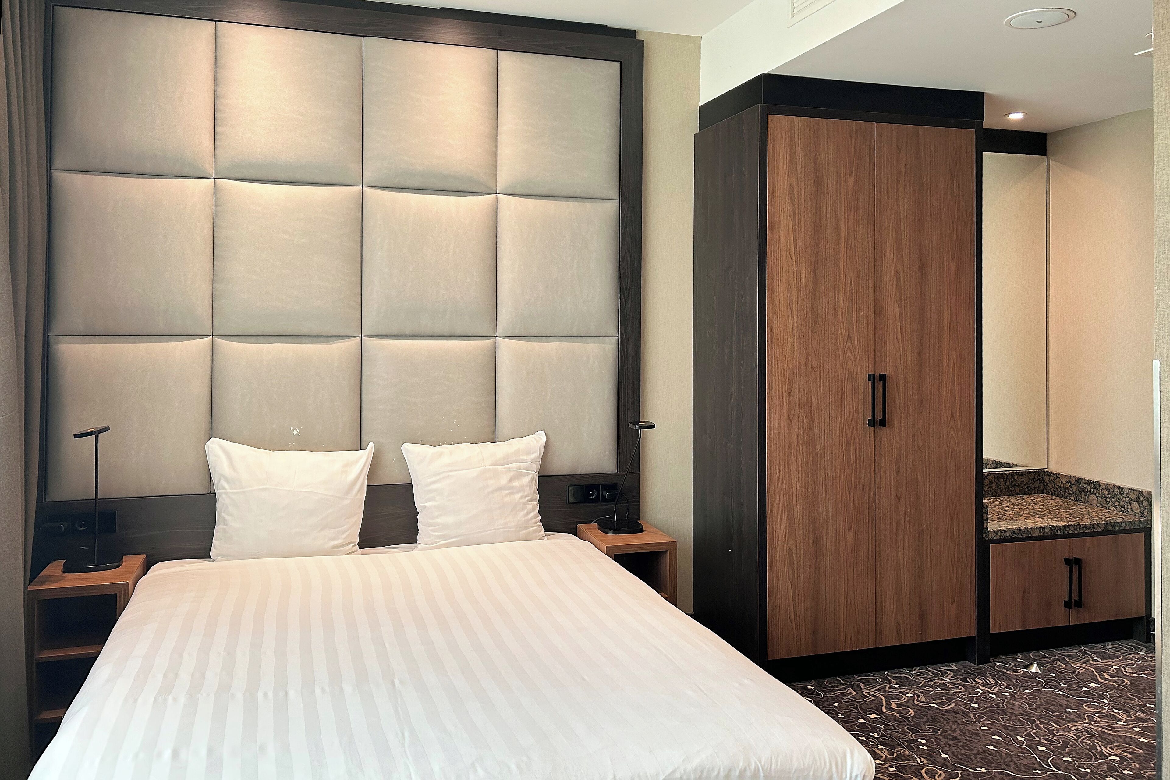 A hotel room with a large bed, a wooden wardrobe, a nightstand, and a mirror.