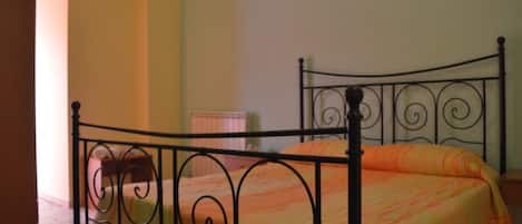 Una cama individual con estructura metálica y colcha a rayas en una habitación con paredes lisas y un radiador.