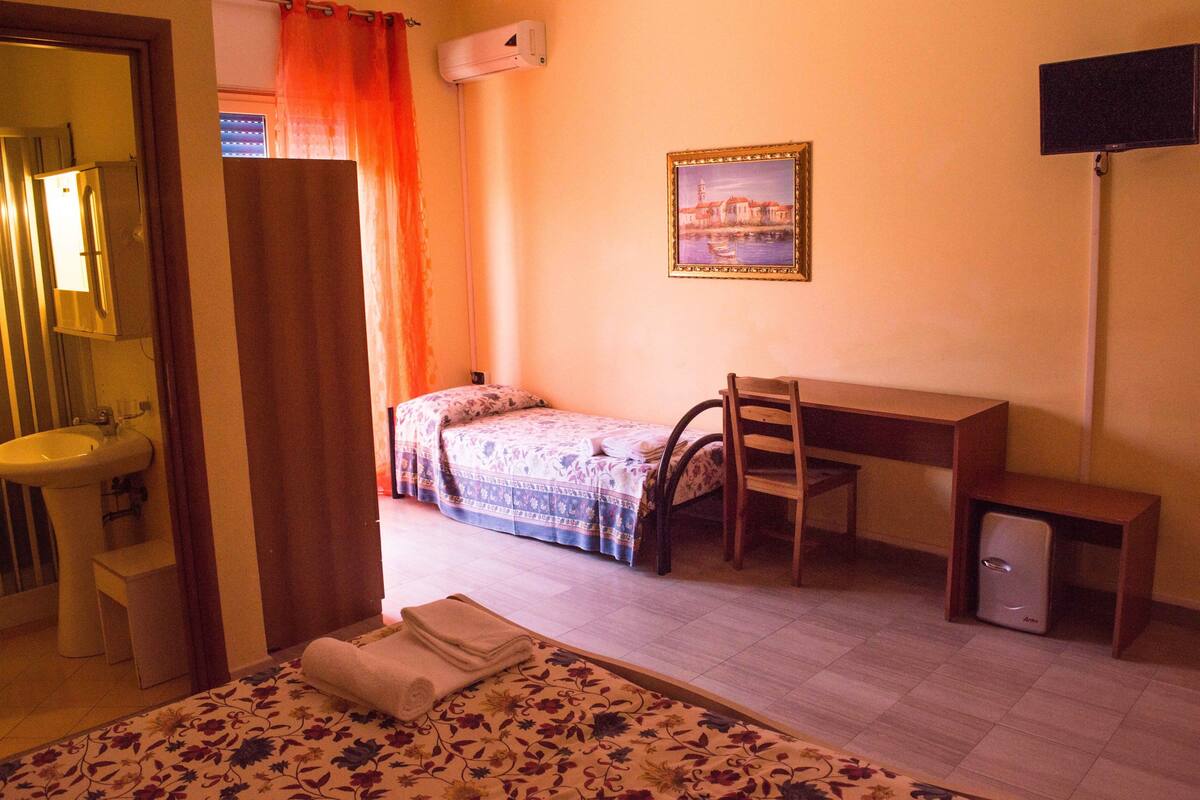 A bedroom with a bed, a desk, a chair, a small cabinet, and a bathroom with a sink and mirror.