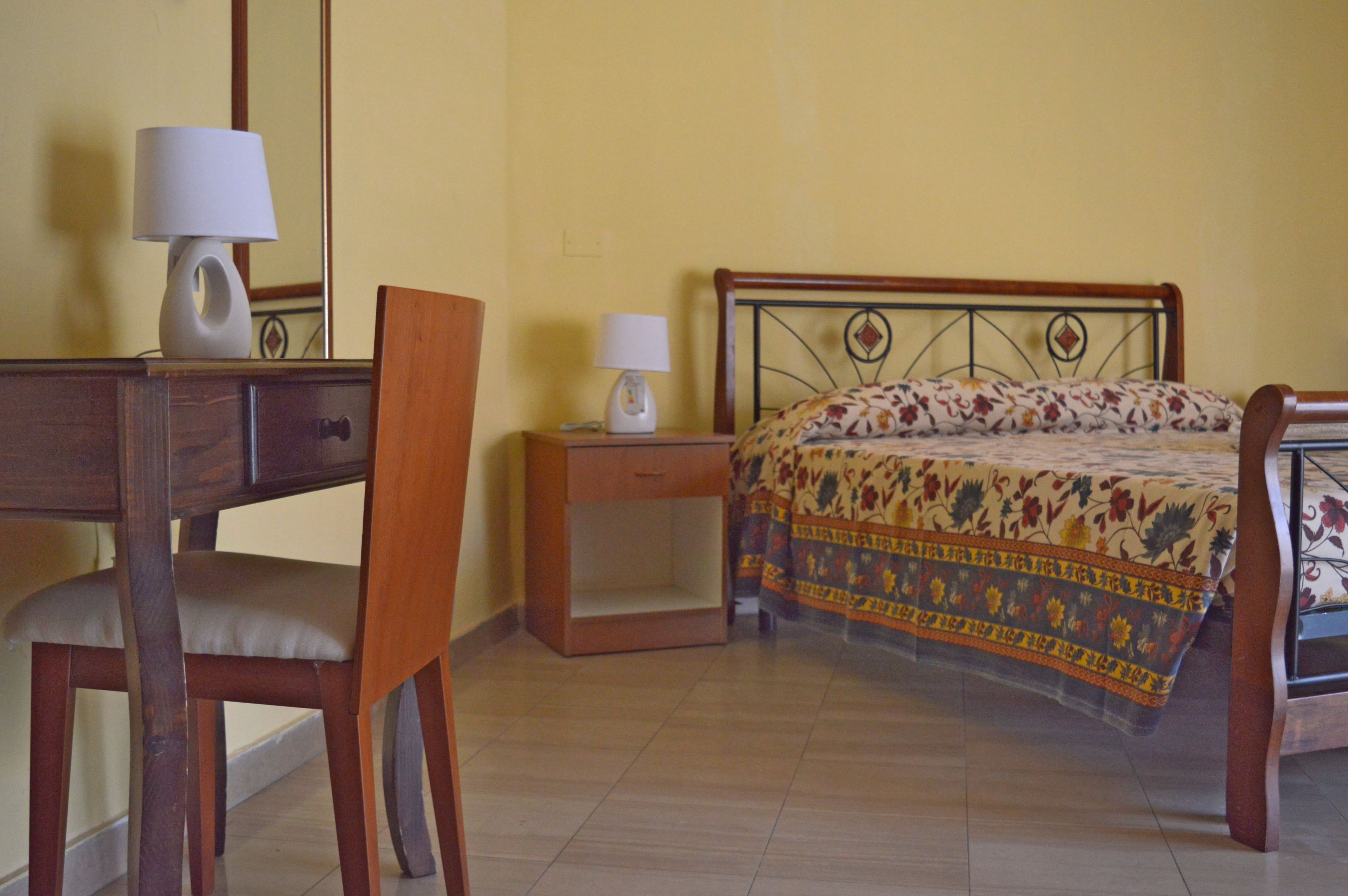 A room with a bed, a desk with a chair, and a nightstand with a lamp.