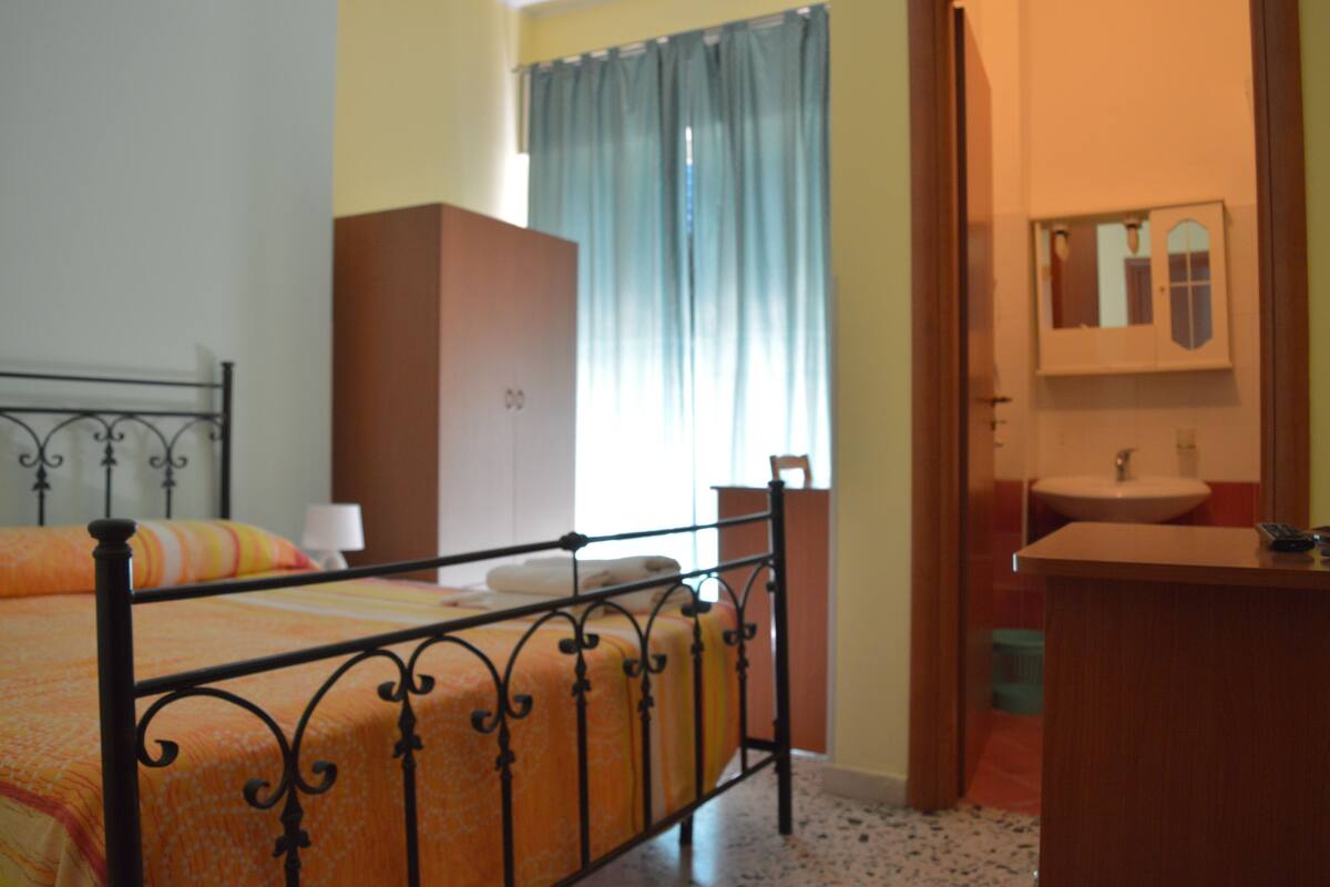 A bedroom with a metal bed frame, a wooden wardrobe, a bathroom with a sink and mirror, and a window with curtains.