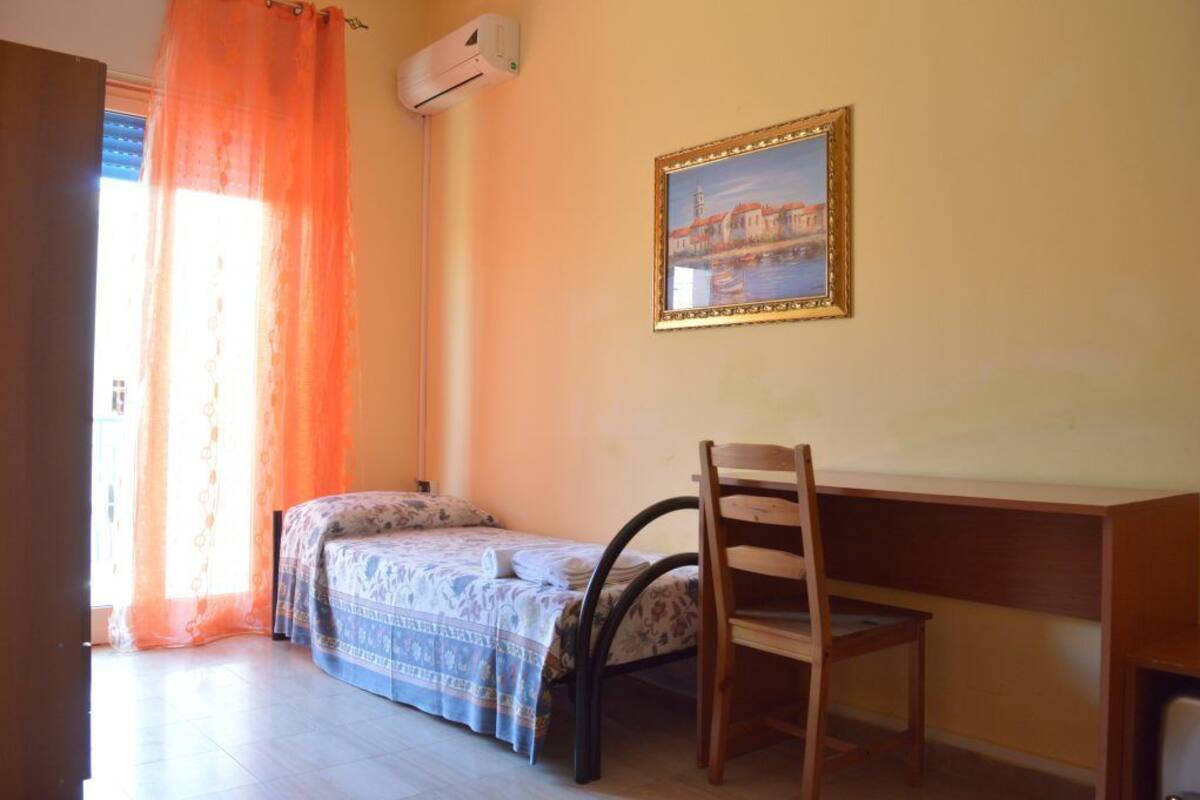 A bedroom with a bed, a desk, and a chair. There is a window with orange curtains, a framed picture on the wall, and an air conditioning unit above the window.