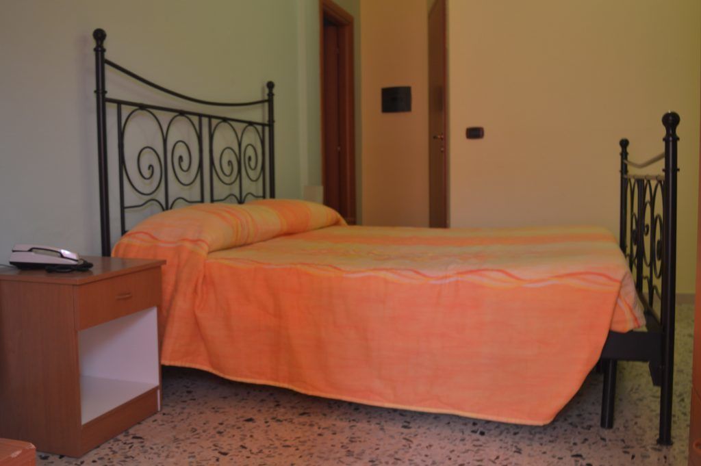 A single bed with an orange bedspread in a room with a speckled floor and a wooden nightstand with a phone.
