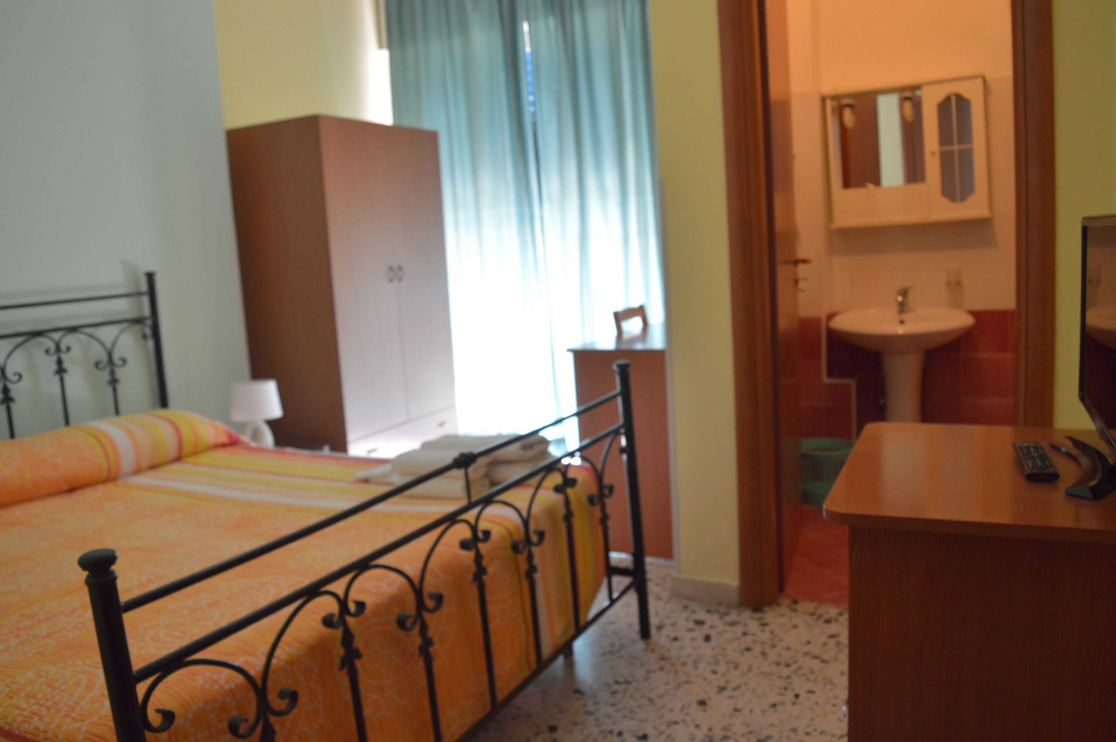 Un dormitorio con estructura de cama de metal, armario de madera, mesita de noche con lámpara y baño con lavabo y espejo.