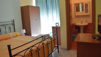 Un dormitorio con estructura de cama de metal, armario de madera, mesita de noche con lámpara y baño con lavabo y espejo.