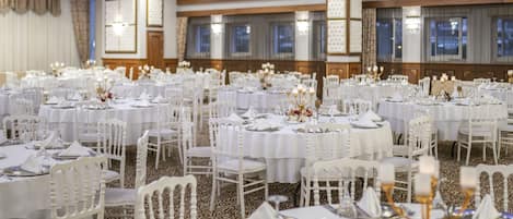 Un salón de banquetes con mesas redondas dispuestas para comer, sillas blancas y candelabros.