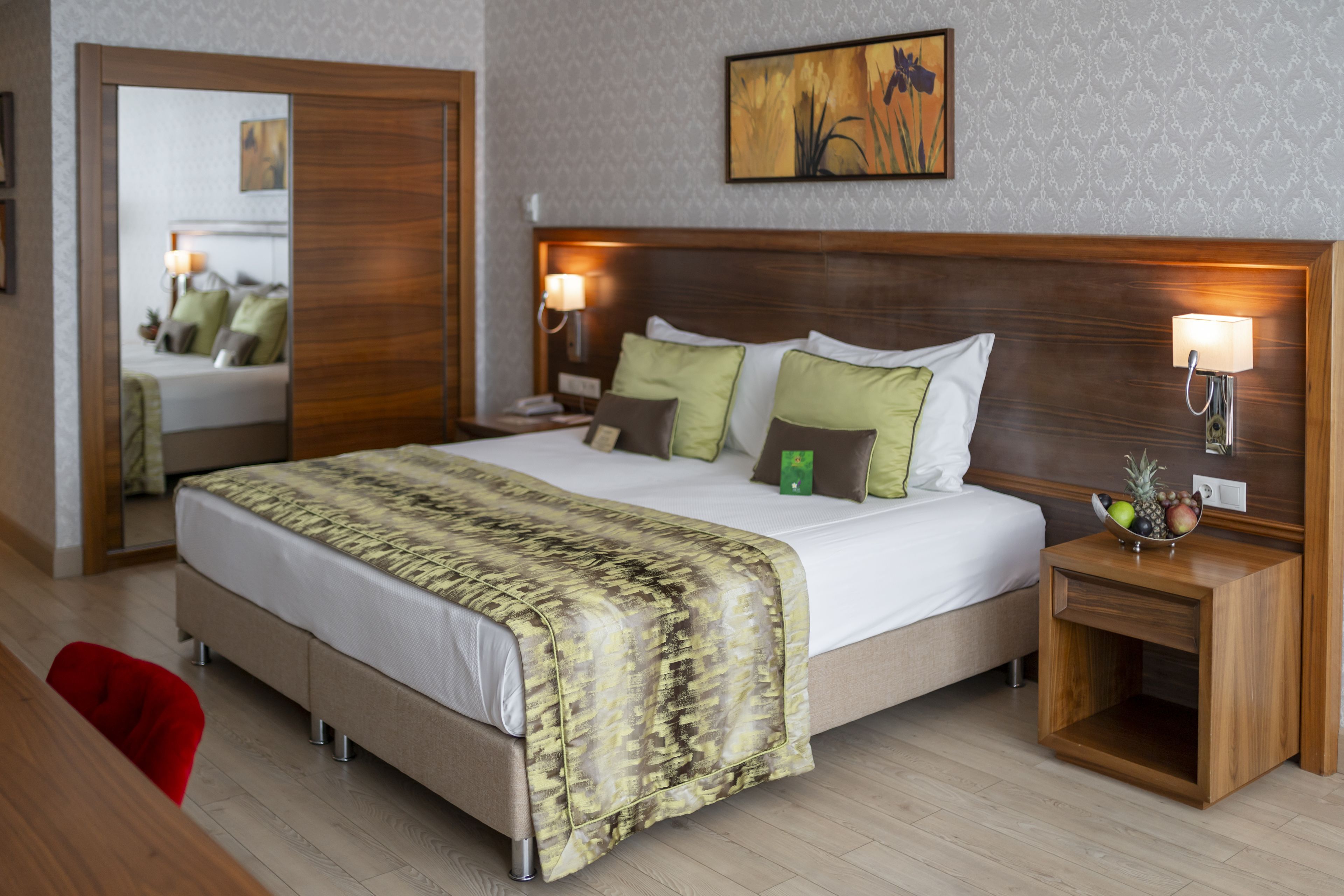 A hotel room with a large bed, wooden headboard, two bedside tables with lamps, a mirror, and a fruit bowl on one table.